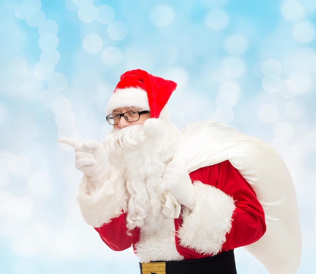 concetto di natale, vacanze e persone - uomo in costume di babbo natale con la borsa che punta il dito su sfondo di luci blu