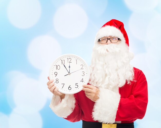 concetto di natale, vacanze e persone - uomo in costume di babbo natale con orologio che mostra dodici su sfondo di luci blu