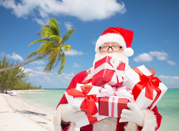 concetto di natale, vacanze e persone - uomo in costume di babbo natale con scatole regalo su sfondo spiaggia tropicale