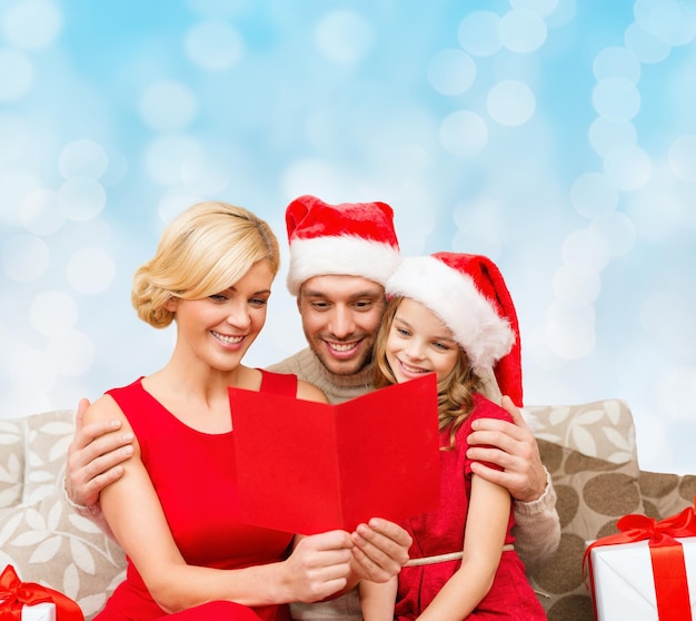 concetto di natale, vacanze, famiglia e persone - felice madre, padre e bambina in cappelli di Babbo Natale con scatole regalo che leggono la carta geeting su sfondo di luci blu