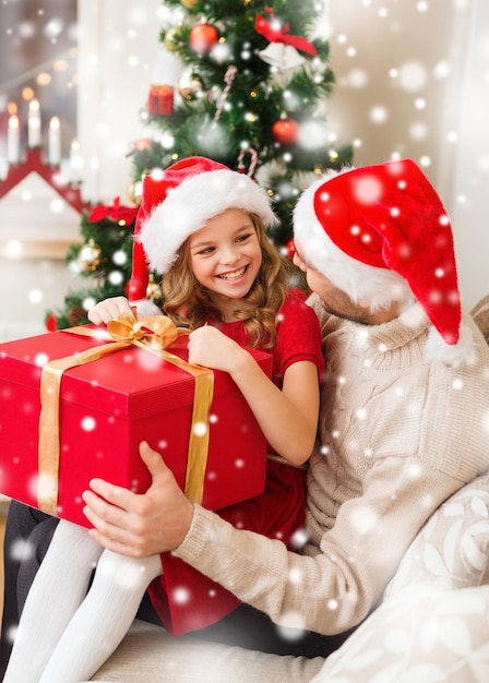 concetto di natale, vacanze, famiglia e persone - padre e figlia sorridenti in cappelli di Babbo Natale che tengono scatola regalo a casa