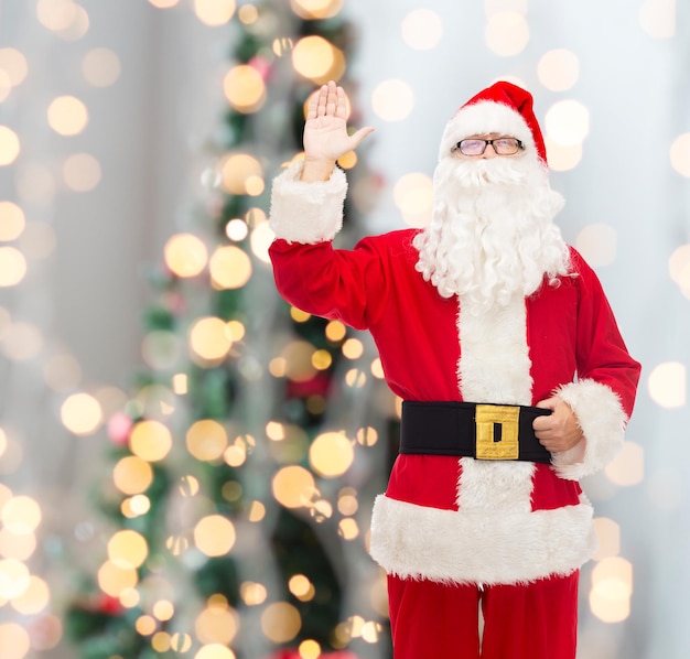 concetto di natale, vacanze, gesto e persone - uomo in costume di babbo natale che agita la mano sullo sfondo delle luci dell'albero