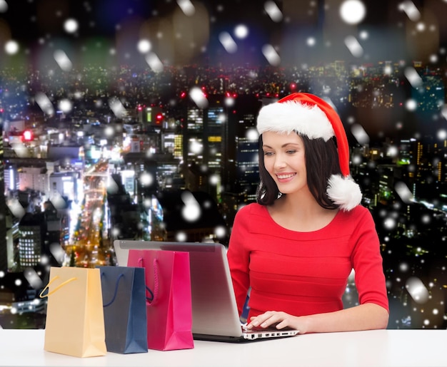 concetto di natale, vacanze, tecnologia e persone - donna sorridente con cappello da Babbo Natale con borse della spesa e computer portatile su sfondo di città di notte innevata