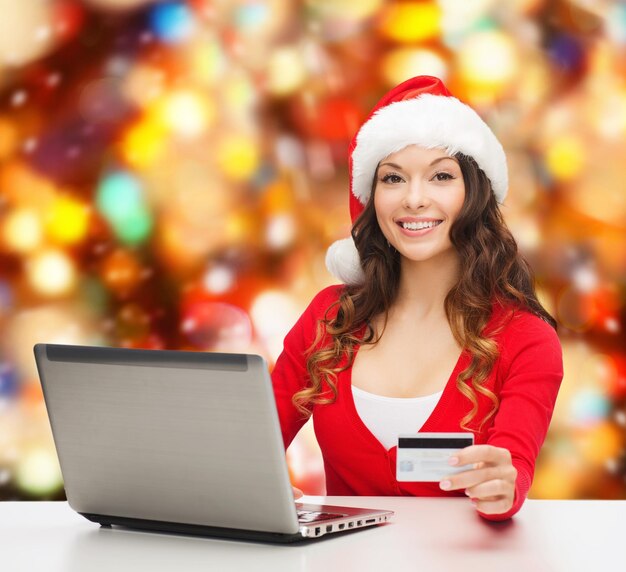 concetto di natale, vacanze, tecnologia e shopping - donna sorridente in cappello di Babbo Natale con carta di credito e computer portatile su sfondo a luci rosse