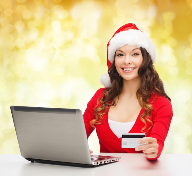 concetto di natale, vacanze, tecnologia e shopping - donna sorridente in cappello di Babbo Natale con carta di credito e computer portatile su sfondo di luci gialle