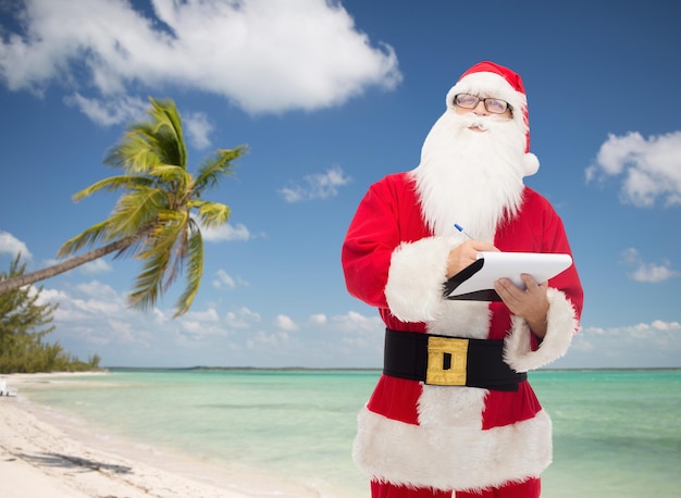 concetto di natale, vacanze, viaggi e persone - uomo in costume di babbo natale con blocco note e penna su sfondo spiaggia tropicale