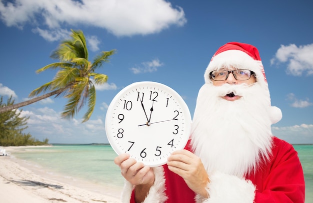 concetto di natale, vacanze, viaggi e persone - uomo in costume di babbo natale con orologio che mostra dodici su sfondo spiaggia tropicale