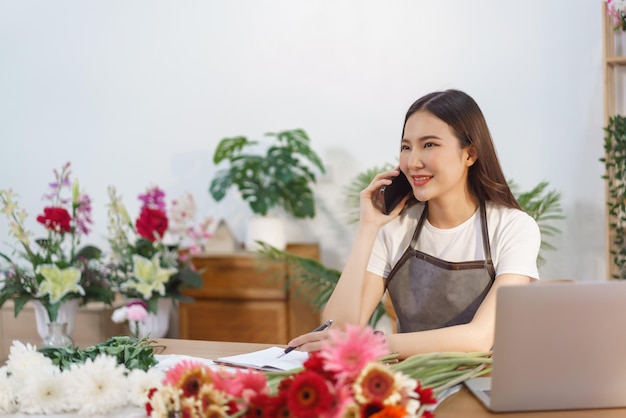 Concetto di negozio di fiori Fiorista femminile che parla con il cliente sullo smartphone e prende nota sul notebook