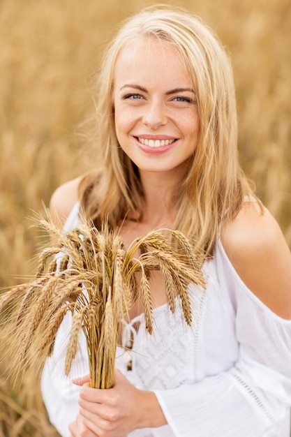 concetto di paese, natura, vacanze estive, vacanza e persone - giovane donna sorridente in abito bianco con spighette che cammina lungo il campo di cereali