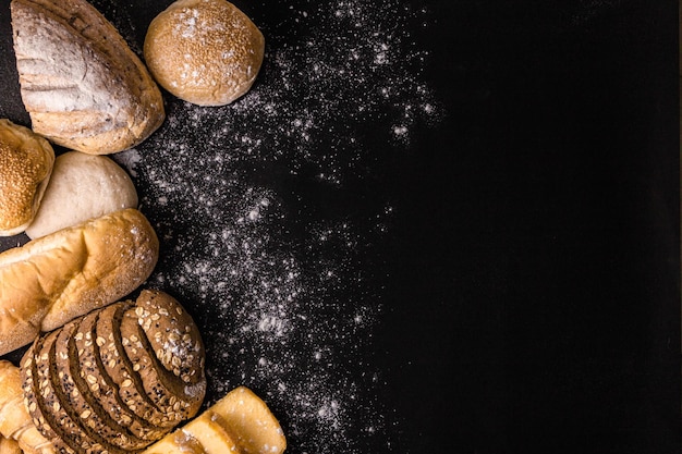 Concetto di pane diverse fette di pane marrone scuro con semi, panini, croissant e pagnotte di pane raccolte insieme sullo sfondo nero con polvere di farina spolverata.