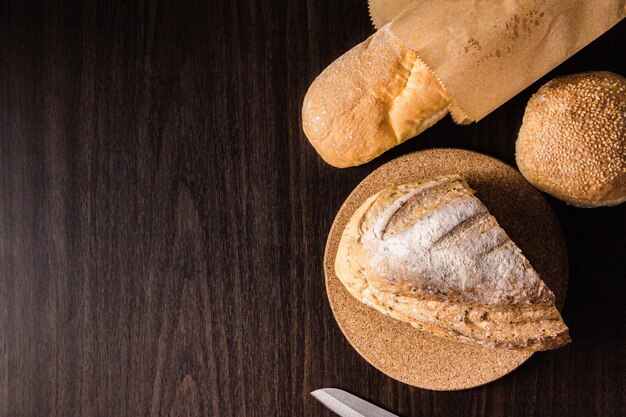 Concetto di pane una mezza pagnotta sul piatto di legno, una pagnotta in un sacchetto di carta, un coltello e un panino messo su una scena di legno nero.