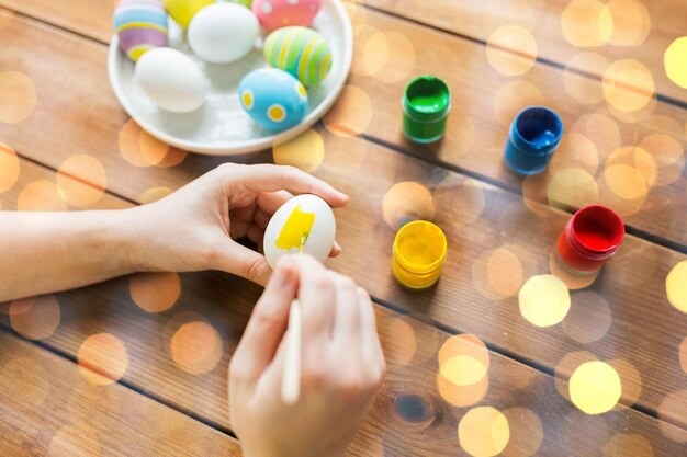 concetto di pasqua, vacanze, tradizione e persone - primo piano delle mani della donna che colorano le uova di pasqua con i colori e spennellano le luci