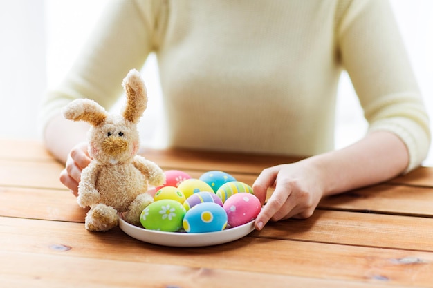 concetto di pasqua, vacanze, tradizione e persone - primo piano delle mani della donna con uova di pasqua colorate su piatto e coniglietto