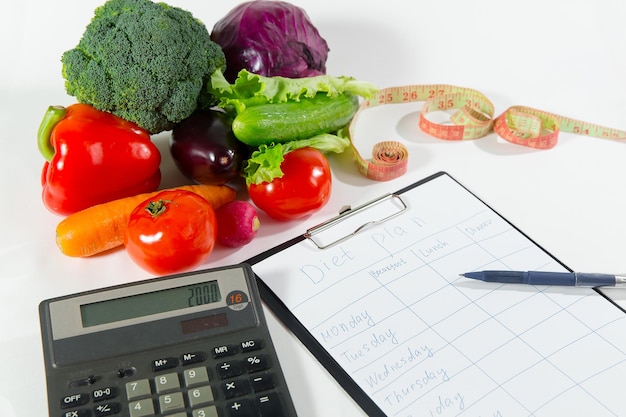 Concetto di perdita di peso, cibo biologico sano. Composizione vegetale, nastro di misurazione, calcolatrice e notebook con piano dietetico isolato su sfondo bianco, vista in primo piano