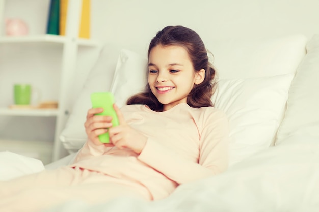 concetto di persone, bambini, riposo e tecnologia - ragazza sorridente felice sdraiata sveglia con lo smartphone a letto a casa