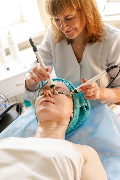 Concetto di persone, bellezza, spa, cosmetologia e tecnologia - Il cosmetologo esegue la procedura di terapia microcorrente della pelle del viso di una bella e giovane donna in un salone di bellezza