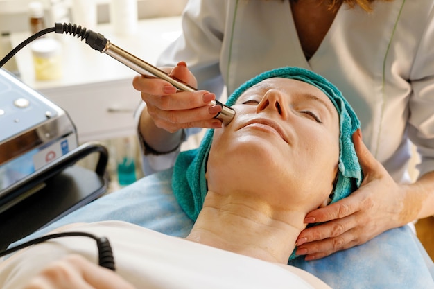 Concetto di persone, bellezza, spa, cosmetologia e tecnologia - Il cosmetologo esegue la procedura di terapia microcorrente della pelle del viso di una bella e giovane donna in un salone di bellezza