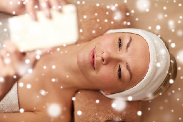 concetto di persone, bellezza, spa, tecnologia e inverno - primo piano di bella giovane donna sorridente sdraiata con lo smartphone nel salone spa con effetto neve