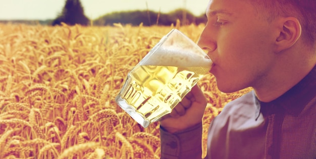 concetto di persone, bevande e alcol - primo piano di un giovane che beve birra da una tazza di vetro su sfondo di campo di cereali