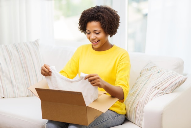 concetto di persone, consegna, commercio, spedizione e servizio postale - giovane donna afroamericana felice che prende i vestiti dalla scatola di cartone o dal pacco a casa