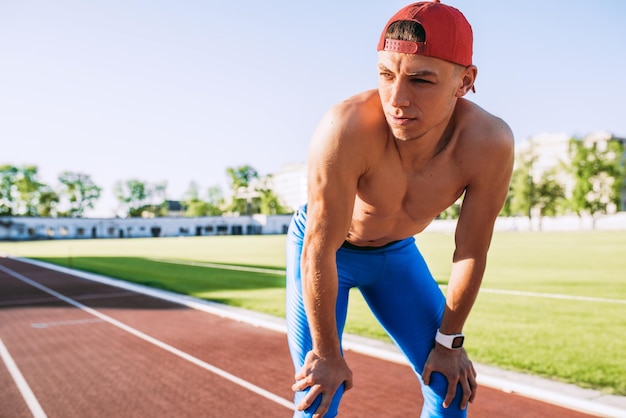 Concetto di persone e sport Primo piano di un bell'atleta maschio che si rilassa in topless dopo un intenso allenamento sullo stadio Runner che si riposa con le mani appoggiate sulle ginocchia dopo un jogging e correre all'aperto