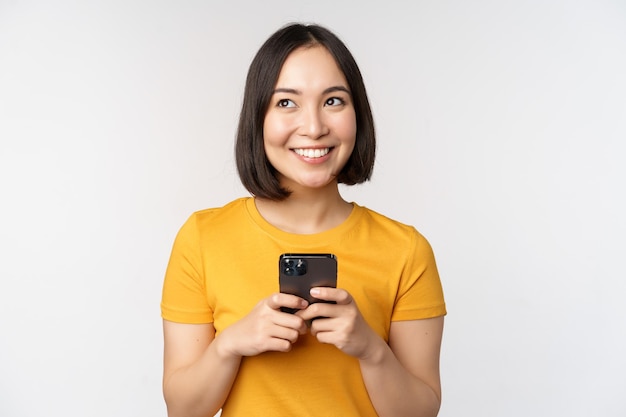 Concetto di persone e tecnologia Ragazza asiatica sorridente che usa messaggi di testo smartphone sul telefono cellulare in piedi su sfondo bianco