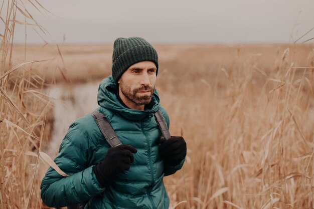 Concetto di persone e turismo Bel giovane pensieroso con la barba lunga con zaino vestito con abiti eleganti concentrato in pose a distanza sullo sfondo del campo all'aperto Stile di vita Avventura