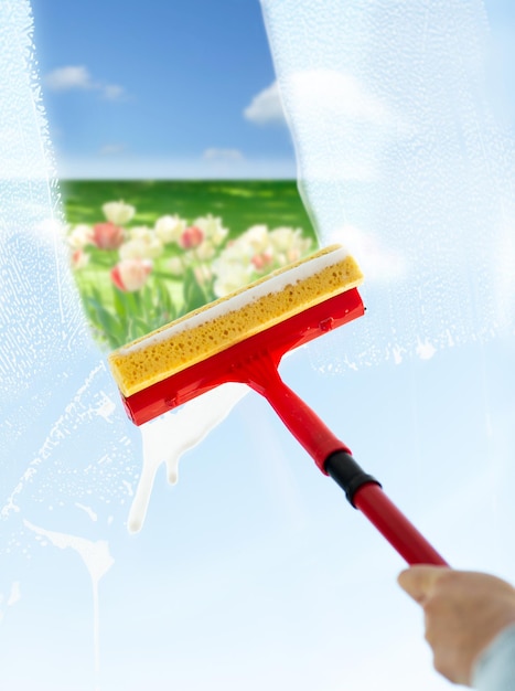 concetto di persone, lavori domestici e pulizie - primo piano del vetro della finestra per la pulizia delle mani con mocio in spugna e schiuma su sfondo blu cielo e campo di fiori