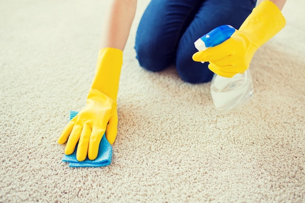 concetto di persone, lavori domestici e pulizie - primo piano di donna in guanti di gomma con panno e detergente spray per la pulizia del tappeto a casa