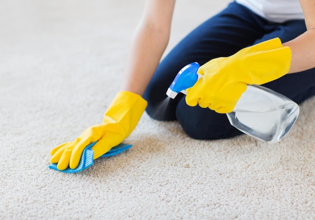 concetto di persone, lavori domestici e pulizie - primo piano di donna in guanti di gomma con panno e detergente spray per la pulizia del tappeto a casa