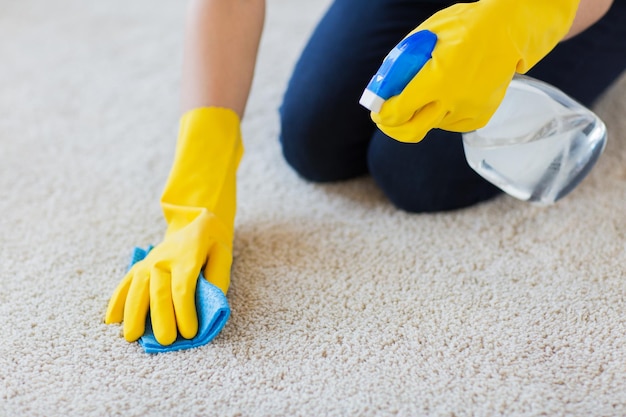 concetto di persone, lavori domestici e pulizie - primo piano di donna in guanti di gomma con panno e detergente spray per la pulizia del tappeto a casa