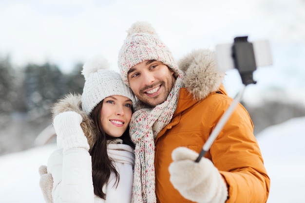 concetto di persone, stagione, amore, tecnologia e tempo libero - coppia felice che scatta foto con lo smartphone su selfie stick su sfondo invernale