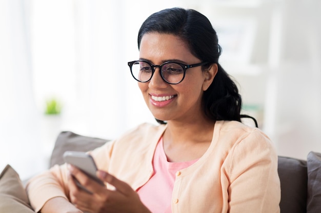 concetto di persone, tecnologia, comunicazione e tempo libero - felice giovane donna indiana con gli occhiali seduta sul divano e messaggio di testo sullo smartphone a casa