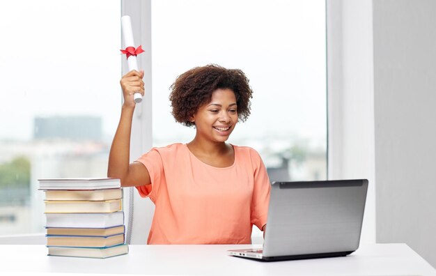 concetto di persone, tecnologia, laurea e istruzione - giovane donna afroamericana felice seduta al tavolo con computer portatile, libri e diploma a casa