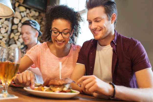 concetto di persone, tempo libero, amicizia, festa e comunicazione - gruppo di amici sorridenti felici che mangiano hamburger insieme al bar o al pub