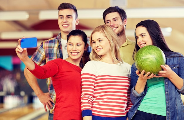 concetto di persone, tempo libero, sport, amicizia e intrattenimento - amici felici che prendono selfie con lo smartphone nel club di bowling