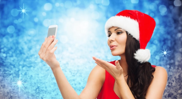 concetto di persone, vacanze, natale e tecnologia - bella donna sexy con cappello rosso da Babbo Natale che scatta foto selfie con lo smartphone e invia un bacio alla fotocamera su sfondo blu glitter o luci