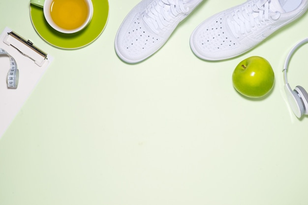 Concetto di piano di fitness. Scarpe da ginnastica, tè, mela e cuffie su sfondo color pastello con taccuino aperto.