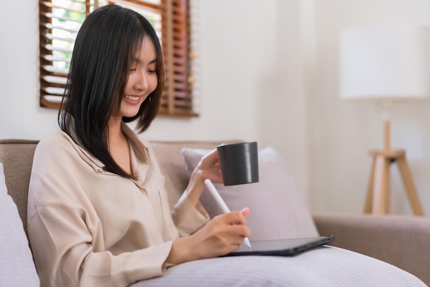 Concetto di relax a casa La donna asiatica sta scrivendo e lavorando sul tablet mentre beve il caffè