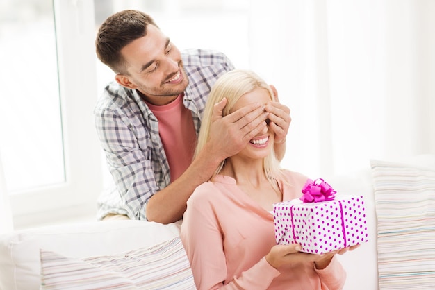 concetto di relazioni, amore, persone, compleanno e vacanze - uomo felice che copre gli occhi della donna e dà una confezione regalo a casa