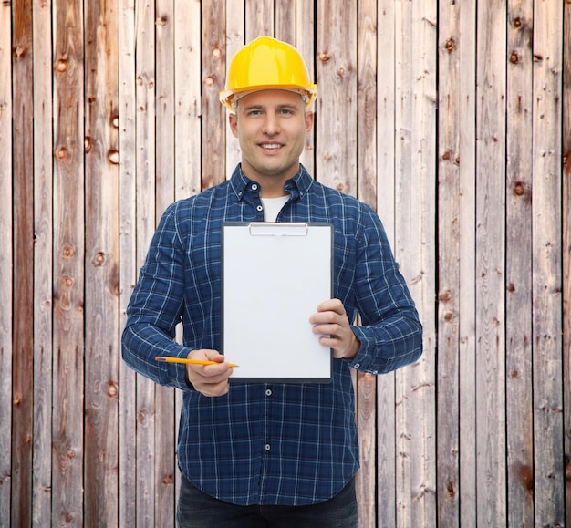 concetto di riparazione, costruzione, costruzione, persone e manutenzione - costruttore maschio sorridente o lavoratore manuale in casco che mostra carta bianca sugli appunti su sfondo di recinzione in legno