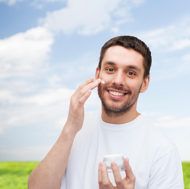 concetto di salute e bellezza - bellissimo uomo sorridente con un vasetto di crema che applica crema