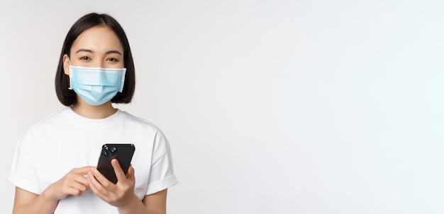 Concetto di salute e covid19 Ragazza asiatica in maschera medica che utilizza l'applicazione per telefono cellulare in quarantena in piedi su sfondo bianco