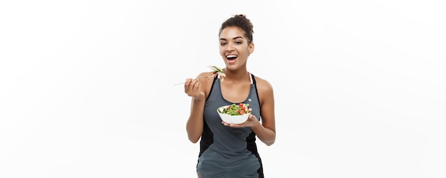 Concetto di salute e fitness Bella signora africana americana in abiti fitness a dieta che mangia insalata fresca isolata su sfondo bianco