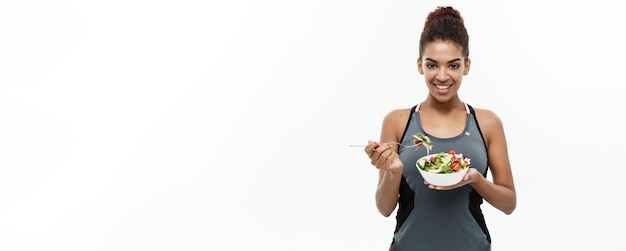Concetto di salute e fitness Bella signora africana americana in abiti fitness a dieta che mangia insalata fresca isolata su sfondo bianco