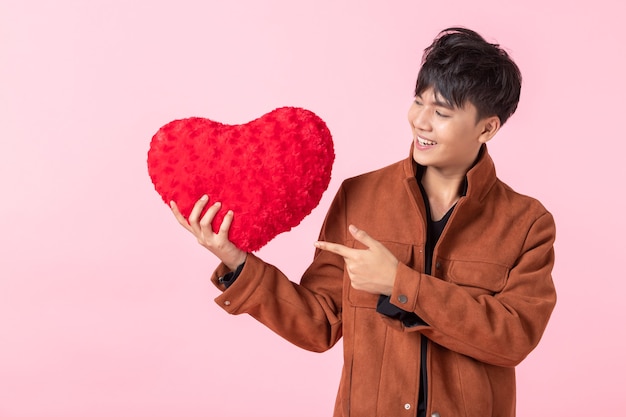 Concetto di San Valentino, asiatico un uomo bello giovane che tiene un cuscino a forma di cuore rosso innamorato isolato su sfondo rosa spazio copia vuoto studio.