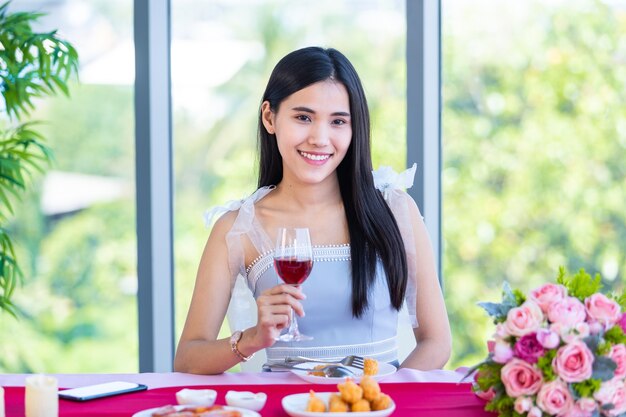 Concetto di San Valentino, ragazza asiatica seduta a un tavolo cibo con bicchieri di vino e bouquet di rose rosse e rosa vino e aspettando il suo uomo sullo sfondo del ristorante