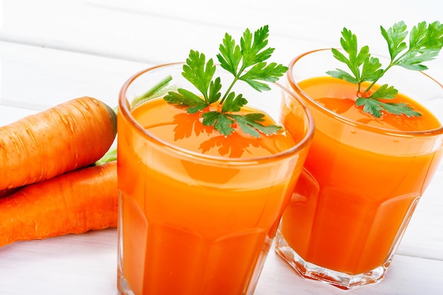 Concetto di sano cibo fatto in casa due bicchieri di succo di carota fresca con prezzemolo accanto a tuberi di carote intere su un tavolo bianco di legno