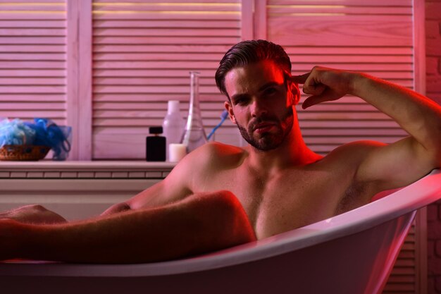 Concetto di sesso ed erotica Macho seduto nudo nella vasca da bagno con luci rosse accese Uomo con barba e faccia seria Ragazzo in bagno con articoli da toeletta sullo sfondo messa a fuoco selettiva