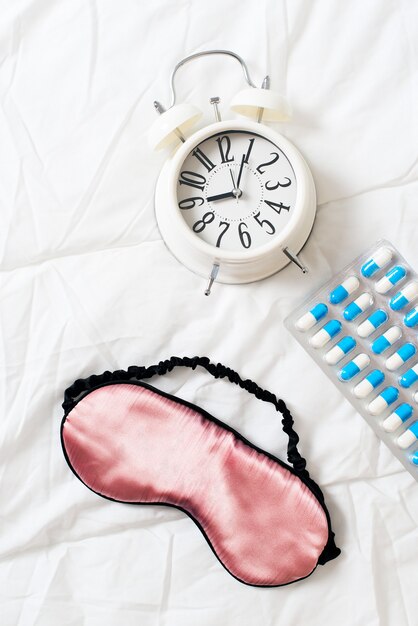 Concetto di sonno sano con maschera, allarme e pillole su vista dall'alto bianco, verticale